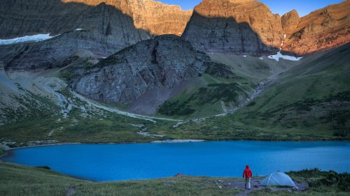 A person in a red jacket stands by a serene blue lake, surrounded by towering mountains at sunrise. A tent is nearby.