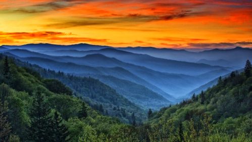 A stunning sunset over layered mountains, with vibrant orange and purple skies and lush green valleys below.