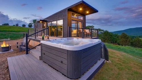 A modern cabin with a hot tub on a deck, surrounded by green hills and a sunset sky. Cozy outdoor seating nearby.