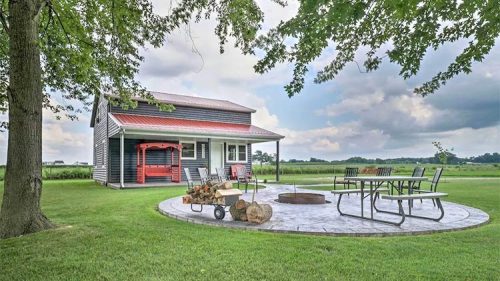 A cozy house with a red porch swing, surrounded by green grass and a circular fire pit area with seating.