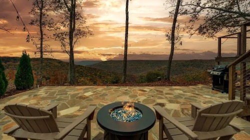 A scenic view of a sunset over mountains, featuring a fire pit and two chairs on a stone patio surrounded by trees.