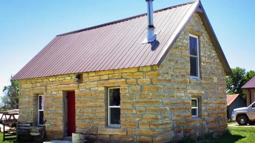 A rustic stone house with a metal roof, featuring a red door and windows, set in a grassy area under a clear blue sky.