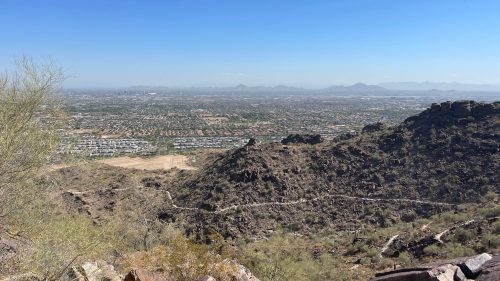 A panoramic view of a desert landscape with rocky hills and a sprawling city in the distance under a clear blue sky.