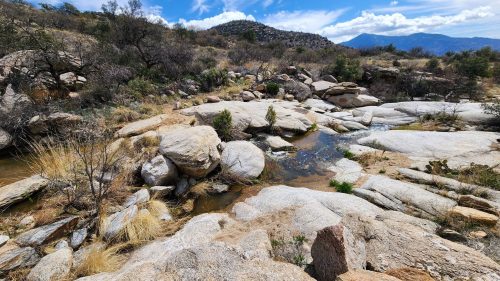 A rocky landscape with scattered boulders, a small stream, and blue skies with wispy clouds in the background.