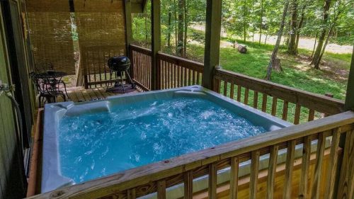 A hot tub on a wooden deck surrounded by trees, with a grill nearby and a peaceful outdoor view.