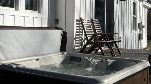 A hot tub with bubbling water in the foreground, and two wooden chairs on a deck beside a white house.
