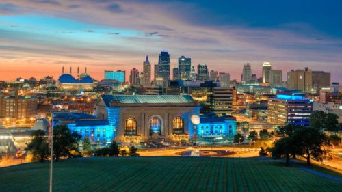A vibrant city skyline at dusk, featuring illuminated buildings and a historic train station in the foreground.