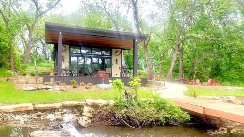 A modern cabin surrounded by greenery, with a small pond and a wooden bridge leading to it.