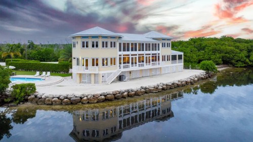 A modern waterfront house with a pool, surrounded by lush greenery and reflecting in calm water under a colorful sky.