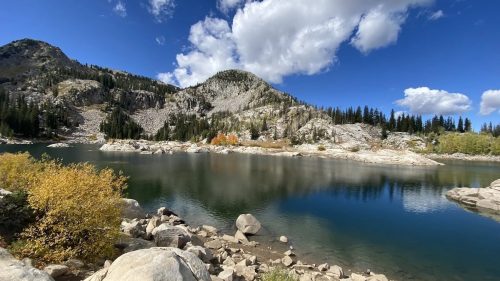 A serene mountain lake surrounded by rocky shores, trees, and vibrant autumn foliage under a blue sky with fluffy clouds.