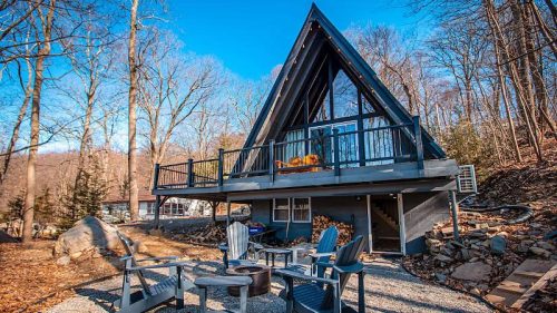 A modern A-frame cabin surrounded by trees, featuring a deck and outdoor seating around a fire pit.