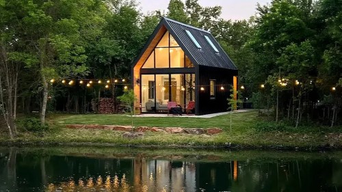 A modern A-frame house by a serene pond, surrounded by trees and illuminated by string lights at dusk.