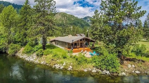 A scenic view of a house by a river, surrounded by trees and mountains under a blue sky with fluffy clouds.