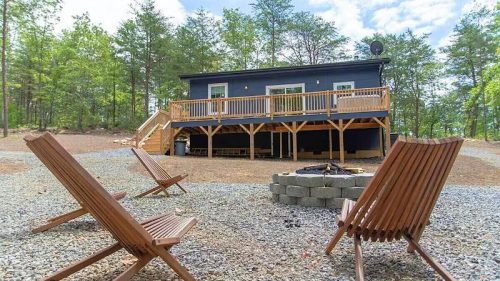 A cozy cabin with a deck surrounded by trees, featuring wooden chairs and a fire pit in the foreground.