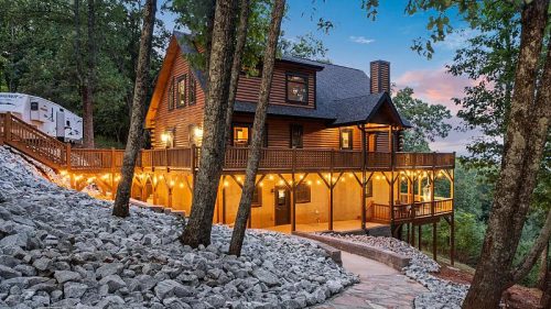 A large wooden house with a wraparound porch, surrounded by trees and a stone pathway leading up to it.