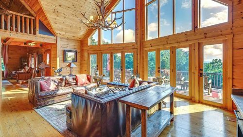 Spacious wooden living room with large windows, a chandelier, and a cozy seating area overlooking a scenic view.