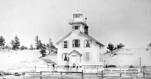 Historic black and white photo of the original Manistee, Michigan, lighthouse and keeper's quarters nestled in sand dunes.