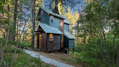 A unique, multi-level cabin nestled among trees, with a wooden pathway leading to its entrance.