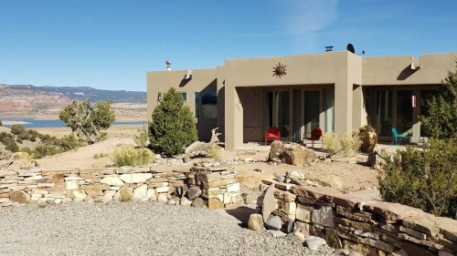 A modern adobe-style house with a sun decoration, surrounded by desert landscape and a view of a lake in the background.