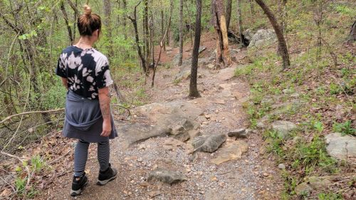 A person walks down a rocky trail surrounded by lush green trees in a forested area.