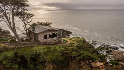 Cozy house on a cliff overlooking the ocean, surrounded by trees and a cloudy sky.