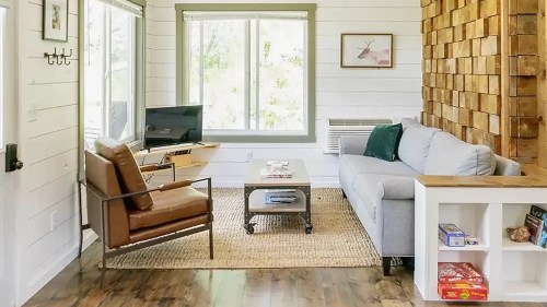 Cozy living room with a gray sofa, wooden accent wall, TV, and a coffee table on a woven rug. Natural light from windows.