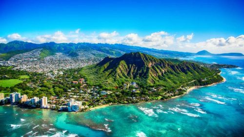 Aerial view of a lush green coastline with mountains, clear blue water, and a vibrant sky.