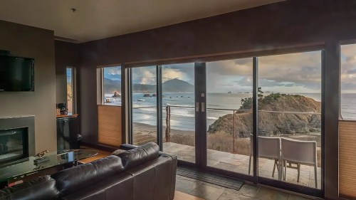A modern living room with a view of the ocean and mountains through large glass doors.