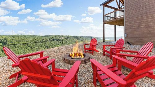 A scenic outdoor fire pit area with red chairs overlooking a lush green landscape and blue sky with clouds.