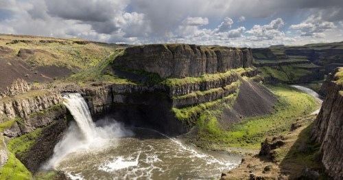A thick waterfall pours out from a rocky outcrop and into a wide basin below. The river flows outward and cuts between tall cliffs that run straight up, seen while on the Palouse Falls Trail in Palouse Falls State Park, Washington.