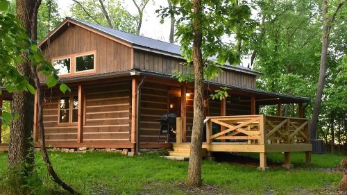 A rustic wooden cabin surrounded by trees, featuring a deck and large windows, set in a lush green landscape.