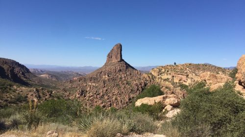 A tall, rocky peak rises above a desert landscape under a clear blue sky, surrounded by rugged hills and vegetation.