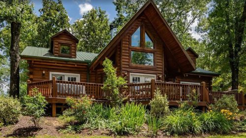 A charming log cabin with a large deck, surrounded by lush greenery and trees under a bright blue sky.