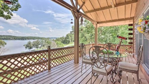 A scenic porch with a table and chairs overlooking a lake and lush greenery under a clear sky.