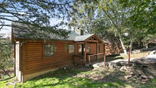 A cozy log cabin surrounded by trees, featuring a front porch and a grassy area.