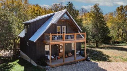 A cozy two-story cabin with a metal roof, surrounded by trees and a gravel path, featuring a welcoming porch.