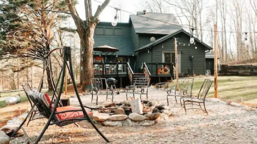 A cozy backyard with a fire pit, seating area, and a dark house surrounded by trees and string lights.