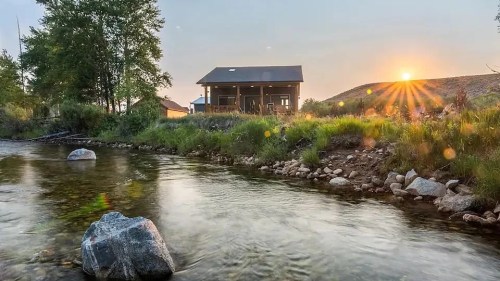 A serene riverside scene at sunset, featuring a house, trees, and rocks along the water's edge.