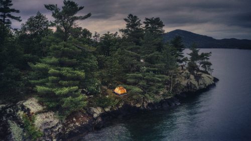 A yellow tent sits on a rocky shore surrounded by trees, with a calm lake and mountains in the background under a cloudy sky.