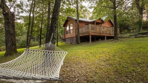 A cozy cabin surrounded by trees, with a hammock in the foreground on a grassy area.