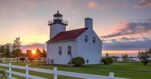 The Sand Point Lighthouse in Escanaba, Michigan, at sunset with a vibrant sunburst and purple clouds.