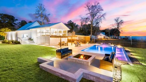 A modern home with a pool and fire pit, surrounded by lush grass, under a colorful sunset sky.