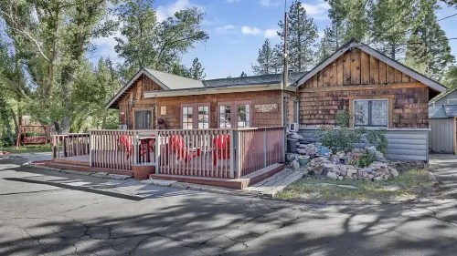 A charming wooden house with a front deck, surrounded by trees and a landscaped yard on a sunny day.