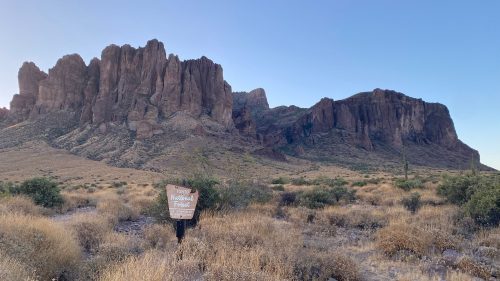 A person holds a sign for a national park in front of rugged mountains and dry, grassy terrain under a clear blue sky.