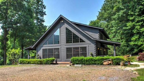 A modern, dark wood house surrounded by trees, featuring large windows and a spacious front porch.