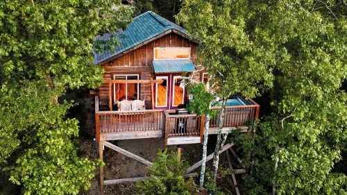 A cozy wooden cabin nestled among lush green trees, featuring a balcony and large windows.