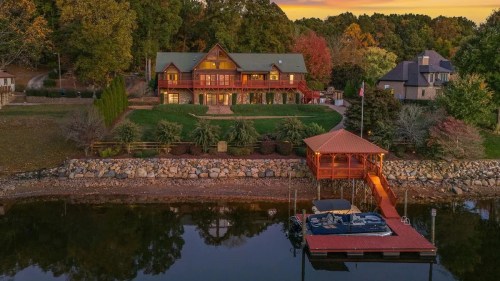 A scenic lakeside view of a large wooden house at sunset, with a dock and colorful autumn trees in the background.