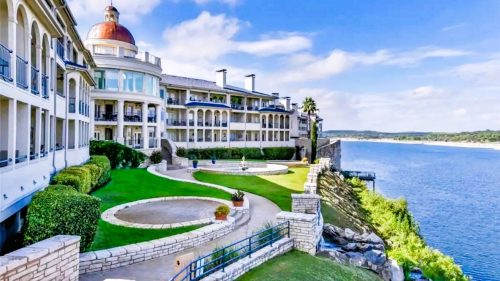 A scenic view of a waterfront hotel with lush green lawns and a blue sky, overlooking a calm lake.