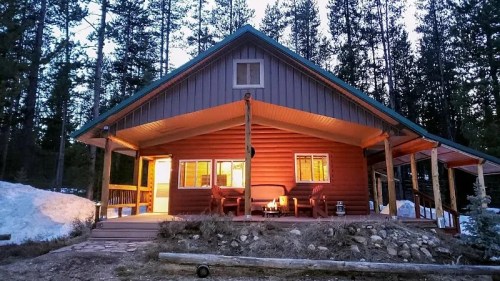 A cozy cabin surrounded by tall trees, with warm light glowing from the windows at dusk. Snow is visible nearby.