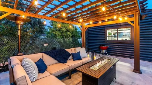 Cozy outdoor patio with a sofa, fire table, and string lights under a wooden pergola, next to a grill and greenery.